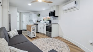 Une cuisine moderne avec des meubles blancs, un four à micro-ondes et un four traditionnel. On y trouve également un ventilateur de plafond, un petit tapis et un canapé gris agrémenté de coussins.