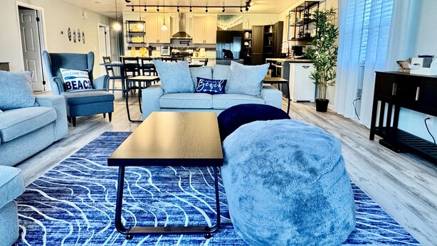 Un salon moderne avec un canapé, un fauteuil et une table basse, un coin repas avec des chaises, et une cuisine en arrière-plan.