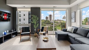 Un salon moderne avec un canapé, un fauteuil et une table basse. Un téléviseur est fixé au mur, et il y a un balcon avec vue sur la ville.