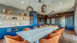 Un espace repas moderne avec une longue table et des chaises orange, un coin cuisine bleu doté d’un four à micro-ondes, et des motifs décoratifs sur les murs.
