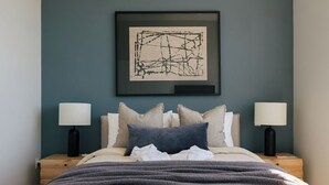 Un lit bien fait, recouvert d’une couverture grise, avec deux lampes de chevet et une œuvre d’art abstraite encadrée accrochée au mur.