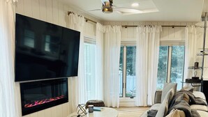 A modern living room with a flat-screen TV mounted on the wall, a round coffee table, a beige sofa, and a fireplace with a glass front.