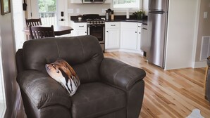 A living room with a leather sofa and a decorative pillow, a dining area with a wooden table and chairs, and a kitchen with white cabinets, a stove, and a refrigerator.