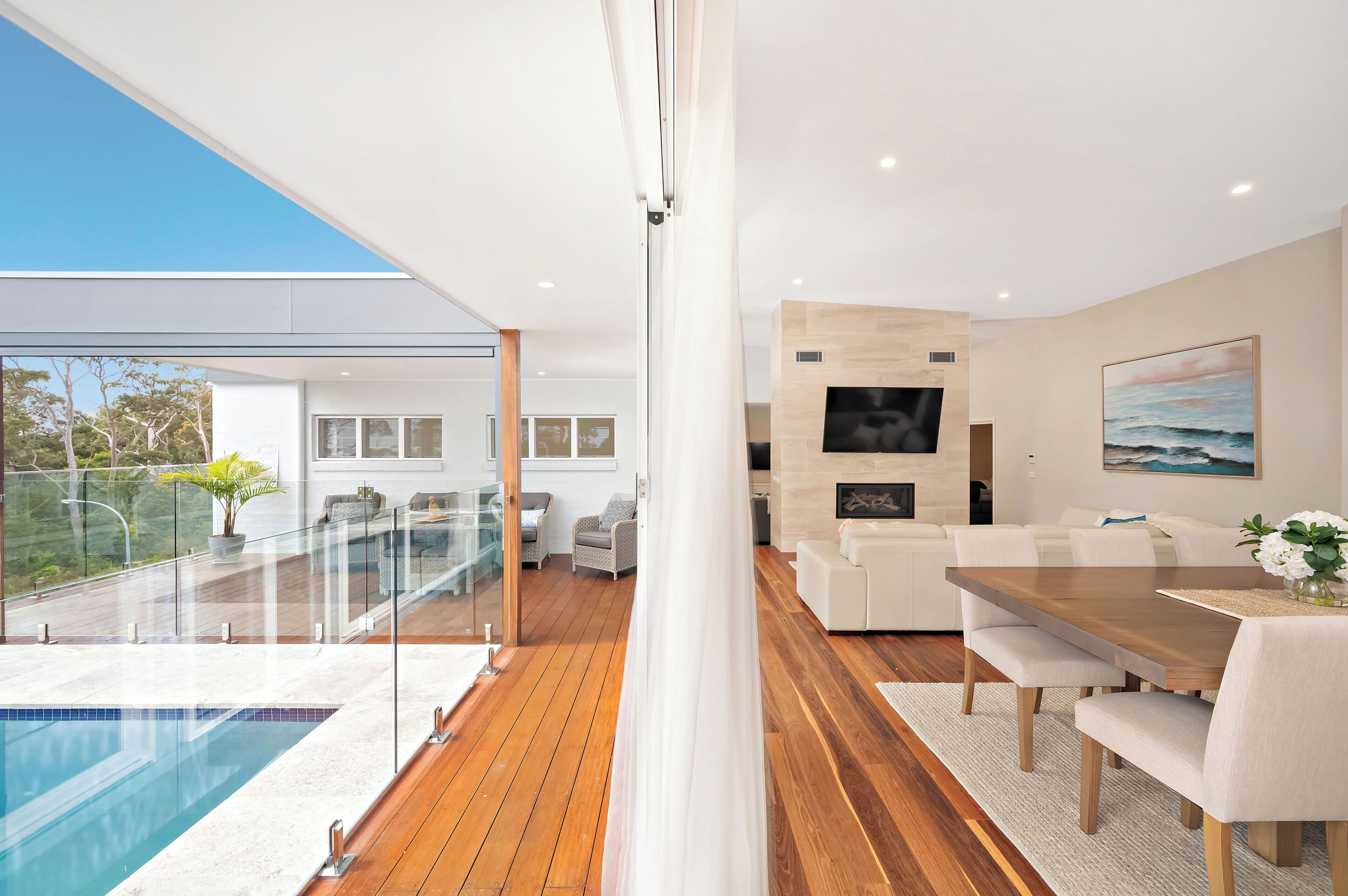 A modern interior with a wooden floor, a dining area with a table and chairs, a living room with a sofa, and a pool area with glass railings.