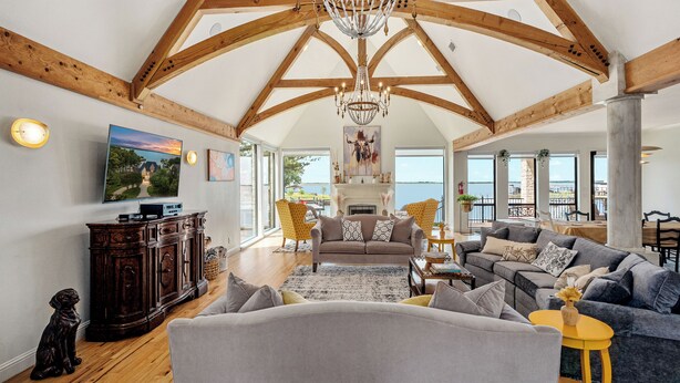 A spacious living room with a high ceiling, wooden beams, a chandelier, and a large window overlooking a scenic view.