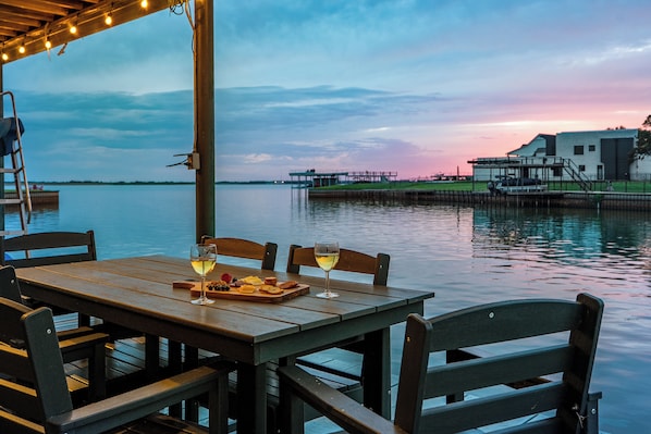 A wooden table with two glasses of wine and a cheese board, overlooking a calm body of water with a dock and buildings in the background.