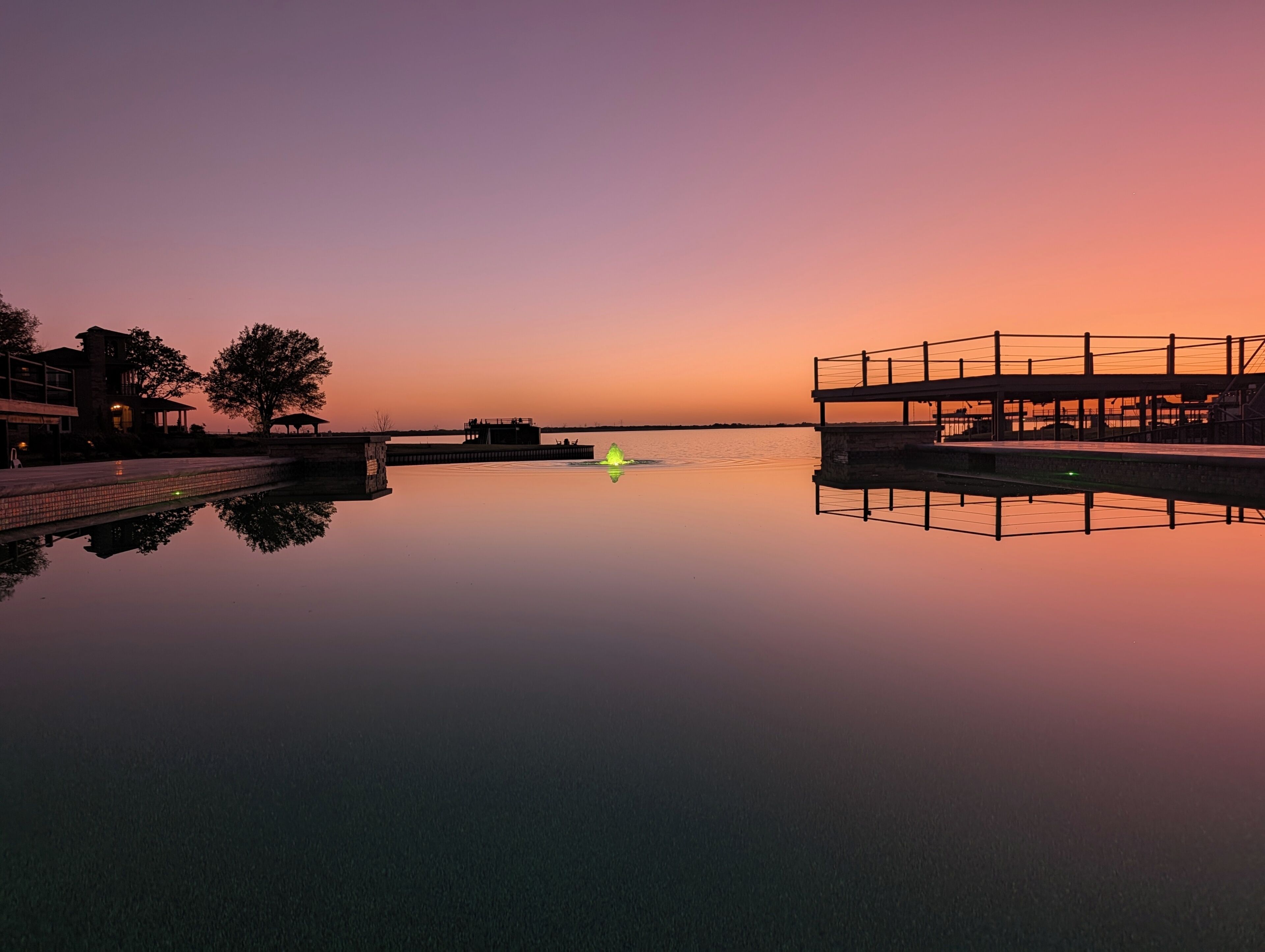 A calm body of water reflecting structures, a dock, and a sunset sky.