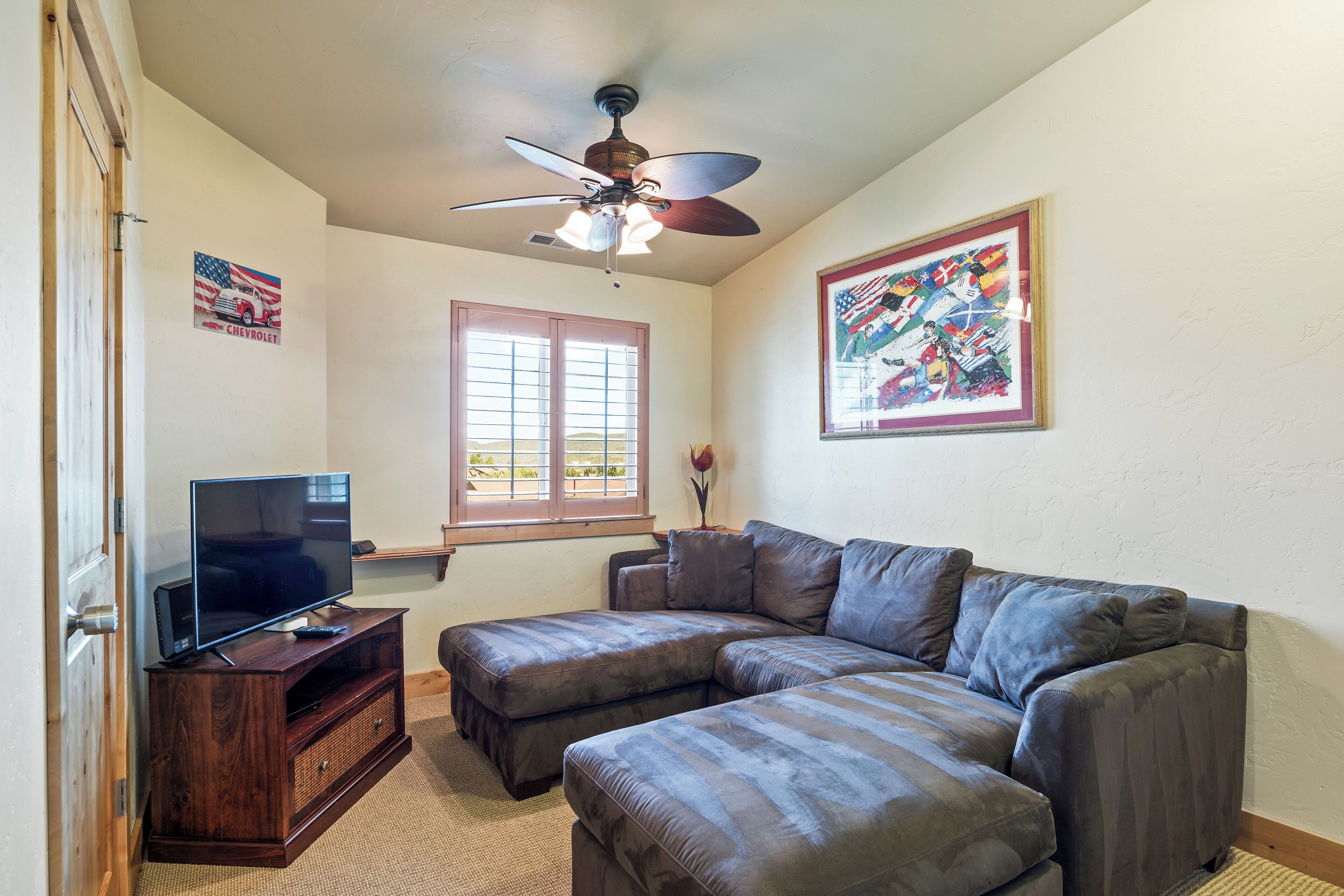 A living room with a sectional sofa, a TV on a wooden stand, a ceiling fan, a window with blinds, and a framed picture on the wall.