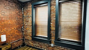 Una habitación con paredes de ladrillo, dos ventanas con marcos oscuros, suelo de madera y una cama con cubierta negra.