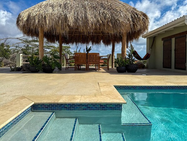 Zona de piscina con una sombrilla de paja, un banco de madera y una piscina con azulejos.