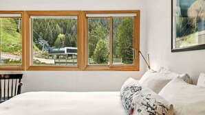 Une chambre avec un grand lit, du linge de lit blanc et une vue sur un télésiège et une forêt, visible à travers trois fenêtres.