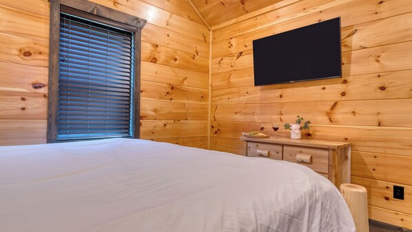 A wooden cabin room with a bed, a dresser, a flat-screen TV, and a window with blinds. - All Fun & Games by Avantstay Indoor Pool, Game Room, Theater, Mountain Views Near Pigeon Forge & Dollywood (Sevierville)