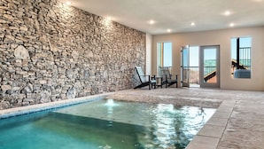Pared de piedra, piscina interior, sillas y una escalera que lleva a un balcón.