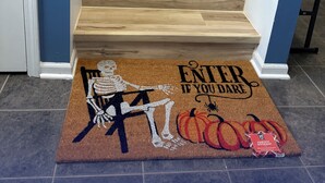Una escalera de madera con un felpudo que muestra un esqueleto, calabazas y la inscripción 'ENTRA SI TE ATREVES'.