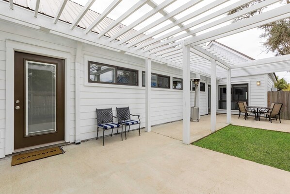 Un patio couvert avec une pergola blanche, une porte dotée d’une fenêtre et une petite pelouse.