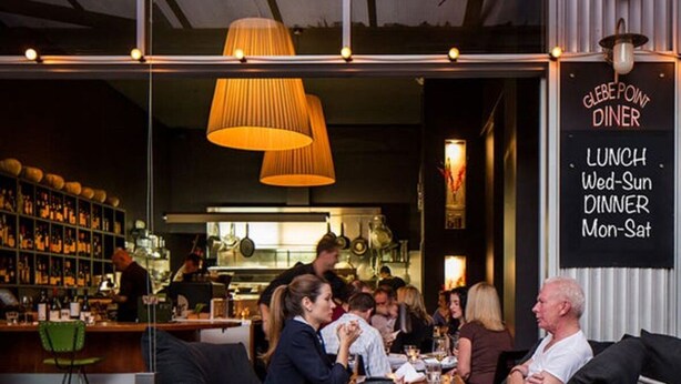 Un espace extérieur aménagé pour les repas, avec des tables et des chaises, un bar doté d’étagères remplies de bouteilles, et un panneau indiquant les heures d’ouverture du restaurant Glebe Point Diner.