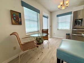 A dining area with a glass table, wicker chairs, and a blue bench. There are windows with blinds, a chandelier, and a stainless steel oven.