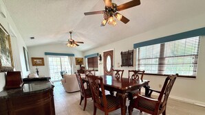 A dining area with a wooden table and chairs, a ceiling fan, and a large window with blinds. - Hammock Beach Cottage (Palm Coast)