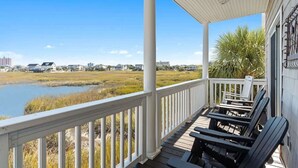 A covered porch with white railings and black Adirondack chairs, overlooking a marshy landscape and a body of water.