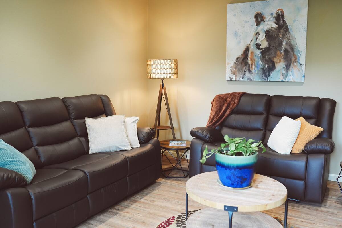 A living room with two dark brown leather sofas, a round wooden coffee table with a blue vase, a floor lamp, and a painting of a dog on the wall.