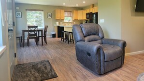 A modern living room with a gray recliner, wooden flooring, and a kitchen in the background. - Sprucewood Dens (Homer)