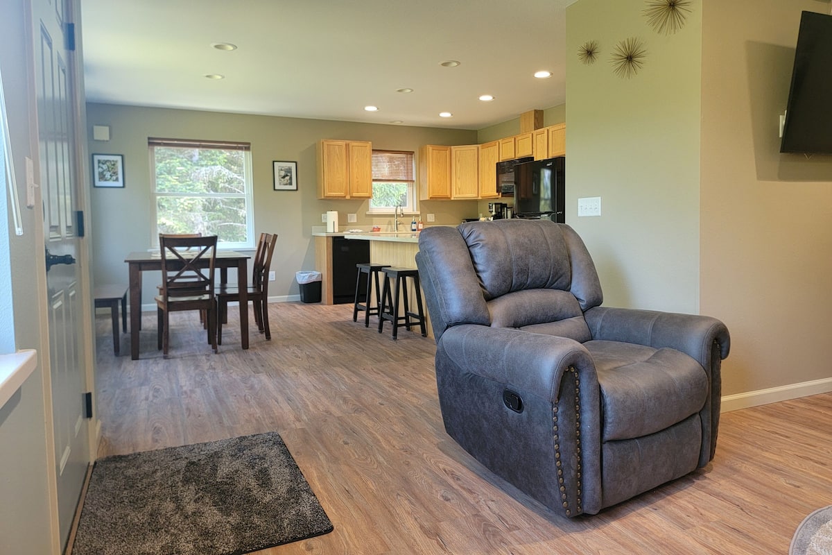 A modern living room with a gray recliner, wooden flooring, and a kitchen in the background.