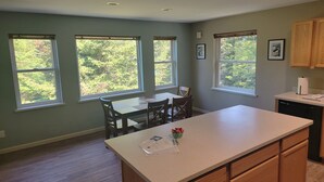 A kitchen with a central island, wooden cabinets, and large windows offering a view of greenery. - Sprucewood Dens (Homer)