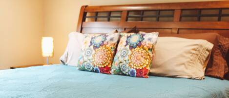 Una cama con un cojín estampado de flores y un cojín liso, un cabecero de madera y una mesita de noche con una lámpara.