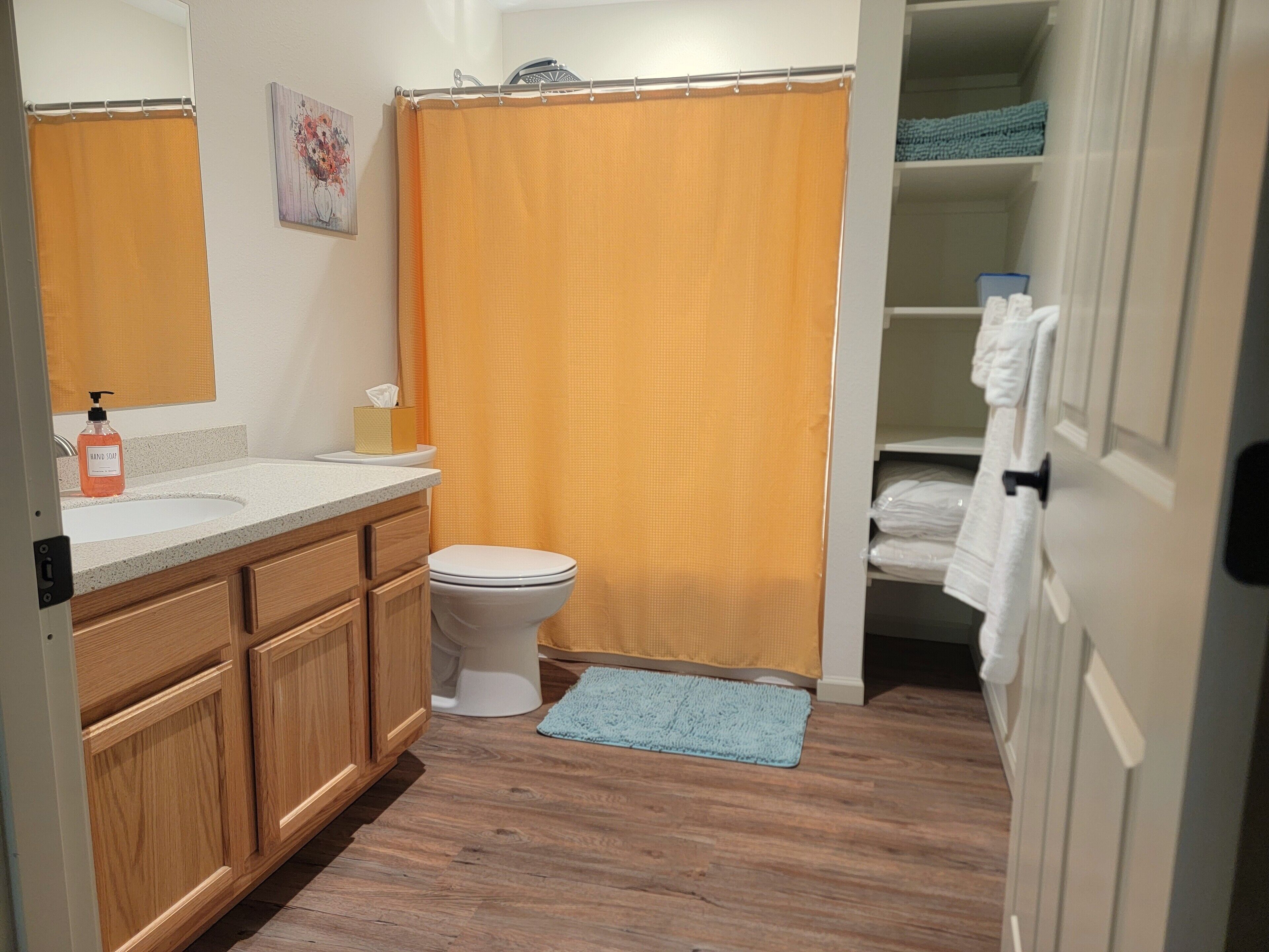 A bathroom with a wooden vanity, a white toilet, an orange shower curtain, and a blue bath mat.