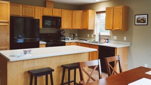 A kitchen with wooden cabinets, a black refrigerator, a microwave, and a coffee maker. There is a window with a view of trees. - Sprucewood Dens (Homer)