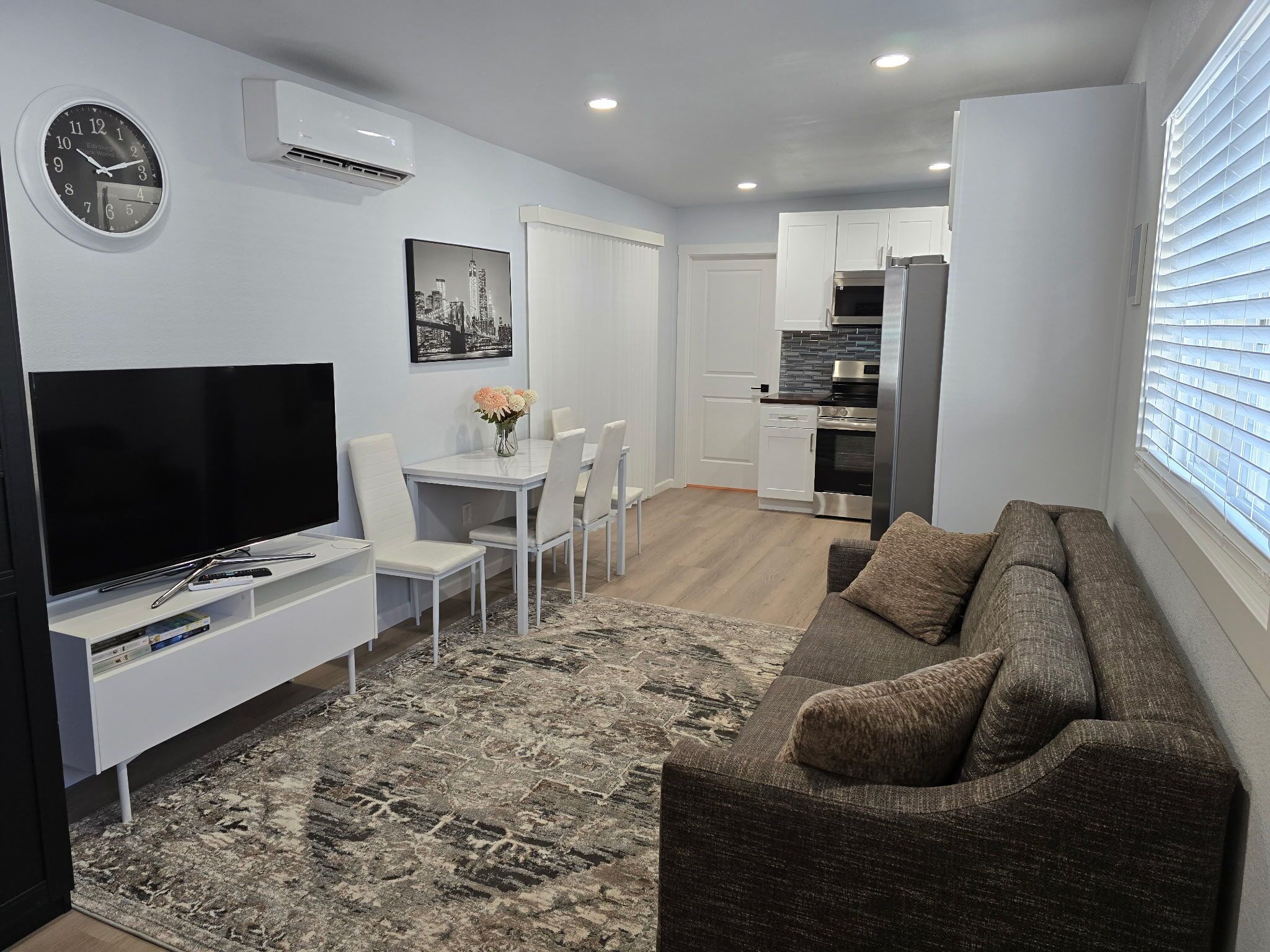 A modern living room with a flat-screen TV, a dining area with a white table and chairs, a gray sofa, and a kitchen with white cabinets and a stainless steel refrigerator.