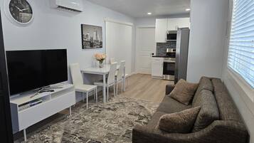 A modern living room with a flat-screen TV, a dining area with a white table and chairs, a gray sofa, and a kitchen with white cabinets and a stainless steel refrigerator.