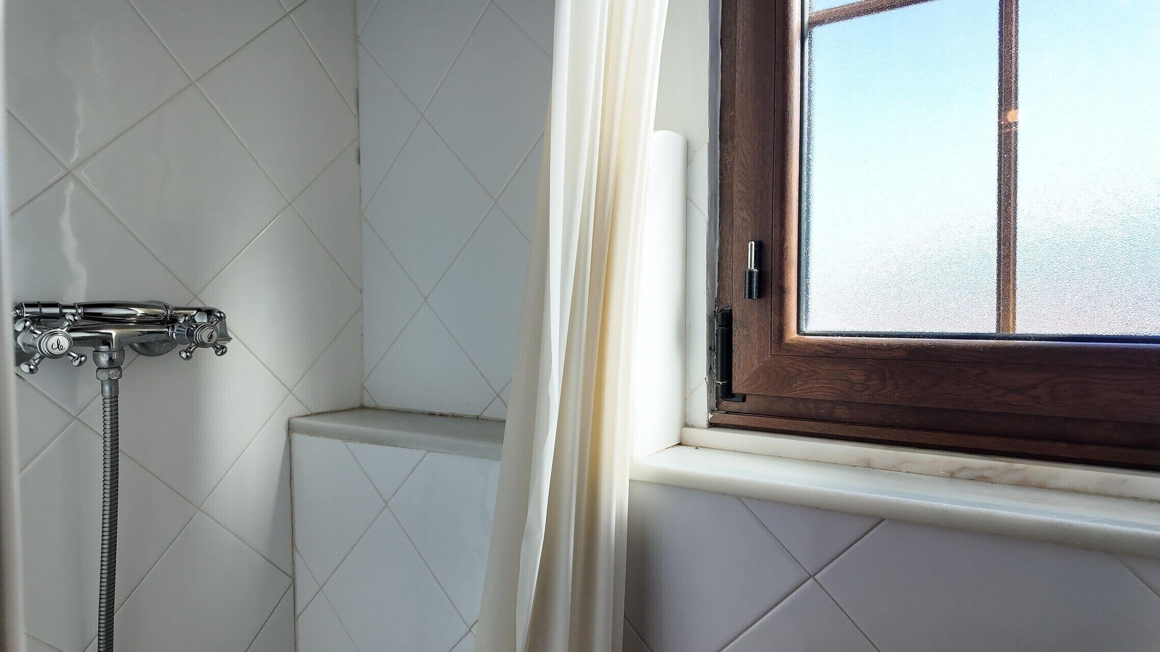 Une salle de bain avec des murs recouverts de carreaux blancs, une douche dotée d’un robinet chromé et une fenêtre avec un cadre en bois.