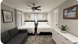 Ein Schlafzimmer mit zwei Betten, einem Deckenventilator, einem weißen Konsolentisch, einem grauen Sofa und einem gemusterten Teppich.