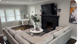 Ein Wohnzimmer mit einem großen, beigefarbenen Ecksofa, einem weißen Couchtisch, einem Kamin, einem Fernseher und einer Topfpflanze.