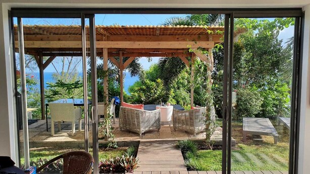 A covered outdoor seating area with wicker furniture and a pergola, overlooking a garden with various plants and trees.