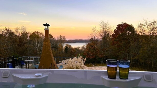 Una vasca idromassaggio con due bicchieri di birra, immersa tra alberi e con vista su un lago al tramonto.