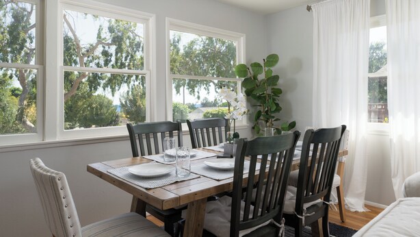 Un coin repas avec une table en bois dressée pour quatre personnes, entourée de chaises, de grandes fenêtres à cadres blancs et une vue sur les arbres à l’extérieur.