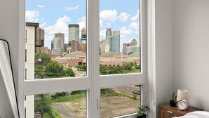A bedroom with a large window offering a cityscape view, a bedside table with a lamp, and a clock. - Luxury 2Bd Condo DT Minneapolis, Minutes From Convention Center & US Bank Arena (Minneapolis)