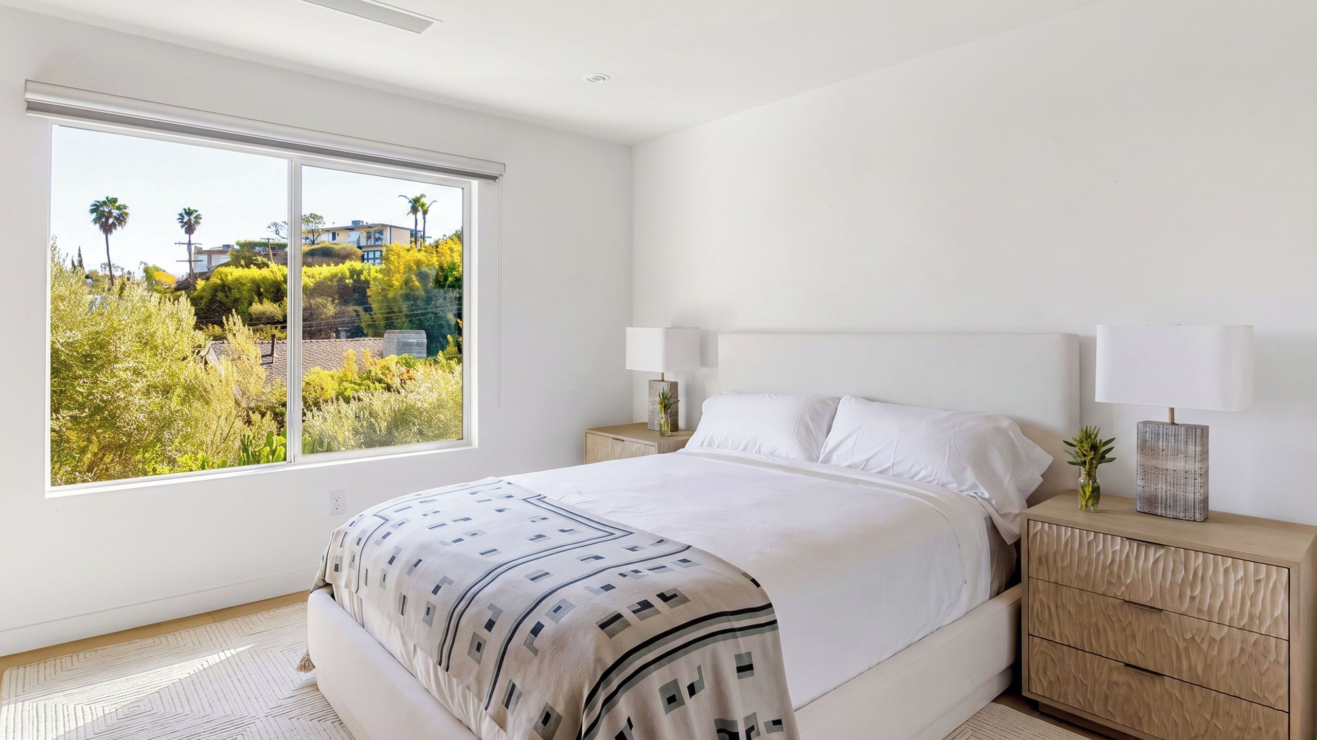 A bedroom with a large window, a bed with a patterned bedspread, two bedside tables with lamps, and a view of greenery outside.