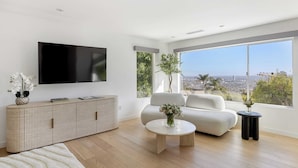 A modern living room with a flat-screen TV, a sofa, a coffee table, and a large window offering a city view. - Luxury Hollywood Hills Home With Pool, Outdoor Dining & Iconic Views Skyline Chalet (West Hollywood)