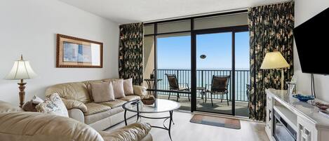 Een woonkamer met een bank, een salontafel, een televisie en een balkon met uitzicht op zee.