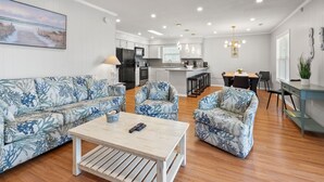 Een moderne woonkamer met een bank, fauteuils en een salontafel. In de achtergrond is een keuken met een koelkast en een eethoek te zien.