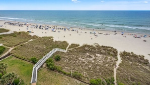A beach with numerous umbrellas and people. There is a white fence and a pathway leading to the beach. - Oceanfront 4BR condo with stunning views, balcony, pool & beach access in North Myrtle Beach (North Myrtle Beach)