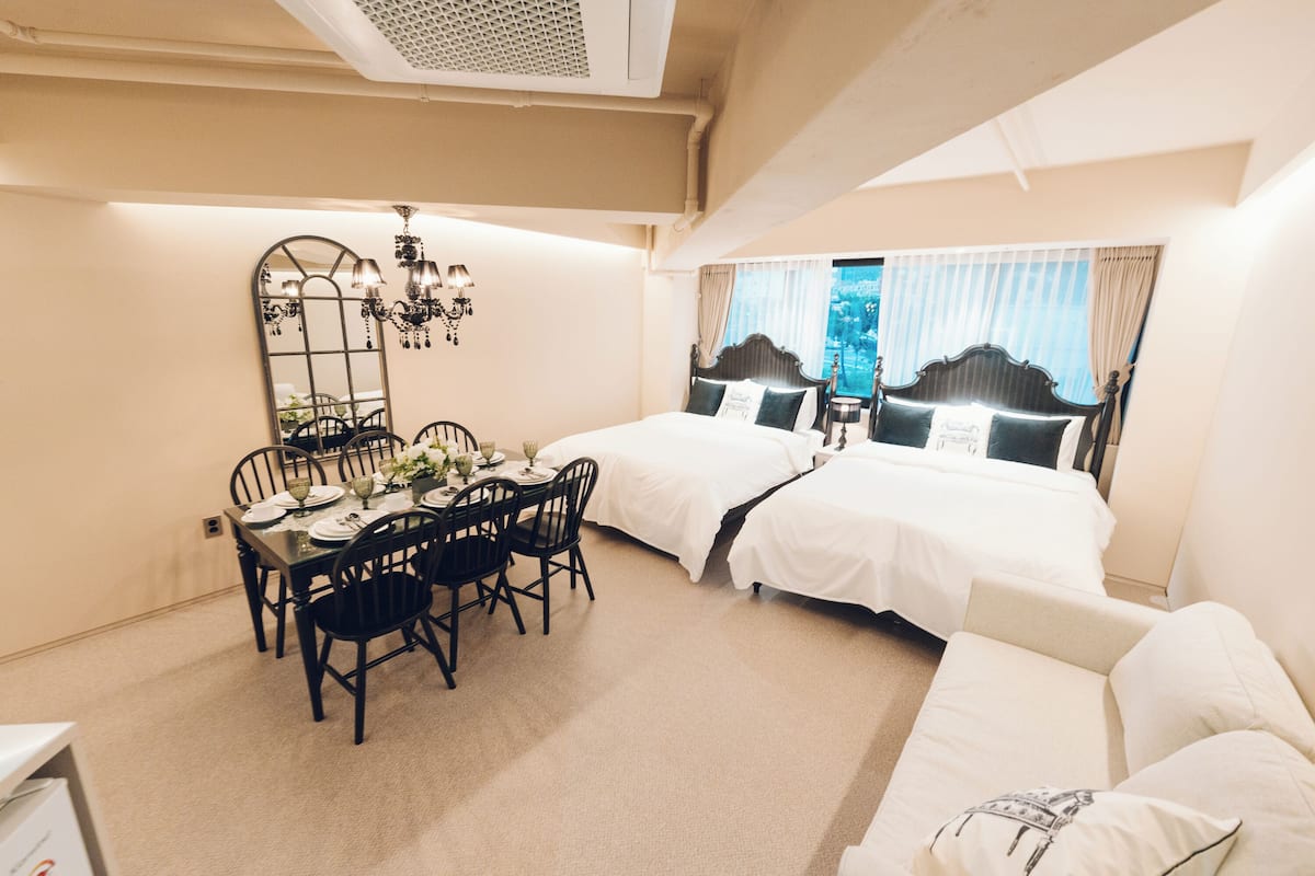 A hotel room with a dining area, two beds, a sofa, a chandelier, and a large mirror.
