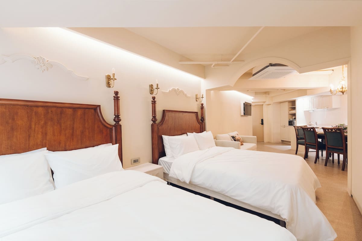 A hotel room with two beds, a wooden headboard, a small table, and a kitchenette in the background.