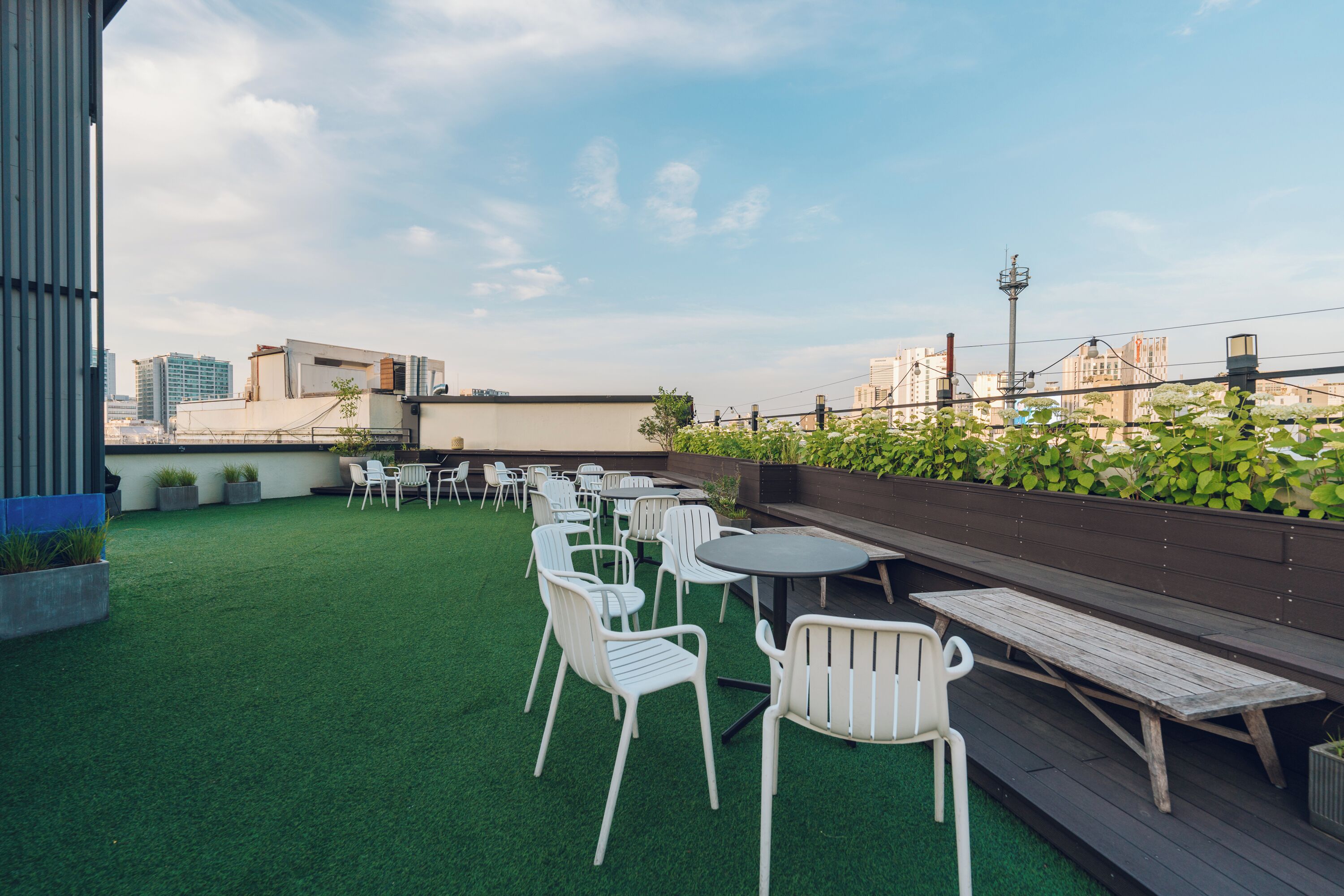 Terraza en la azotea con sillas y mesas blancas, rodeada de plantas verdes y vistas a la ciudad.