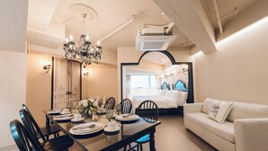 A dining area with a glass table set for four, surrounded by chairs. A chandelier hangs above, and a bed with a blue headboard is visible in the background. - MONTHLIV Euljiro Hotel Stay (Seoul)