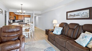 Ein Wohnzimmer mit braunem Ledersofa, einem braunen Ledersessel und einem Esstisch mit weißen Stühlen. Im Hintergrund ist eine Küche mit Holzausbau und einem Kronleuchter zu sehen.
