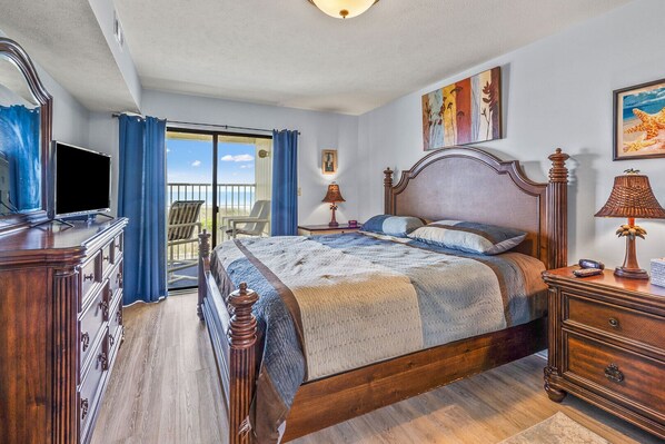 A spacious bedroom with a large bed, a dresser, a television, and a balcony with chairs.